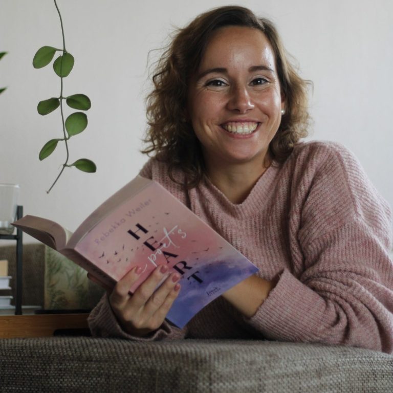 Frau mit lockigem Haar hält ein Buch und lächelt, im Hintergrund Pflanzen sichtbar.