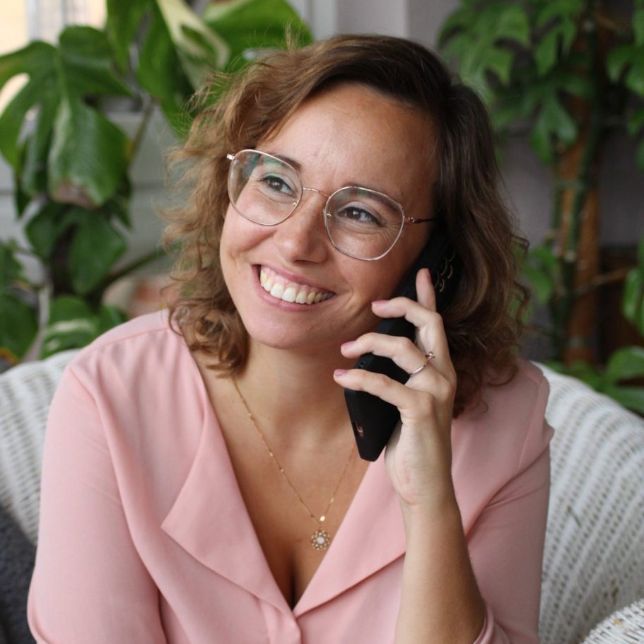 Junge Frau mit Brille lächelt und spricht telefonierend, umgeben von Pflanzen.