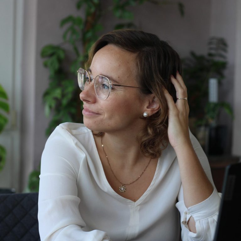 Frau mit Brille sitzt am Tisch und schaut nachdenklich zur Seite.