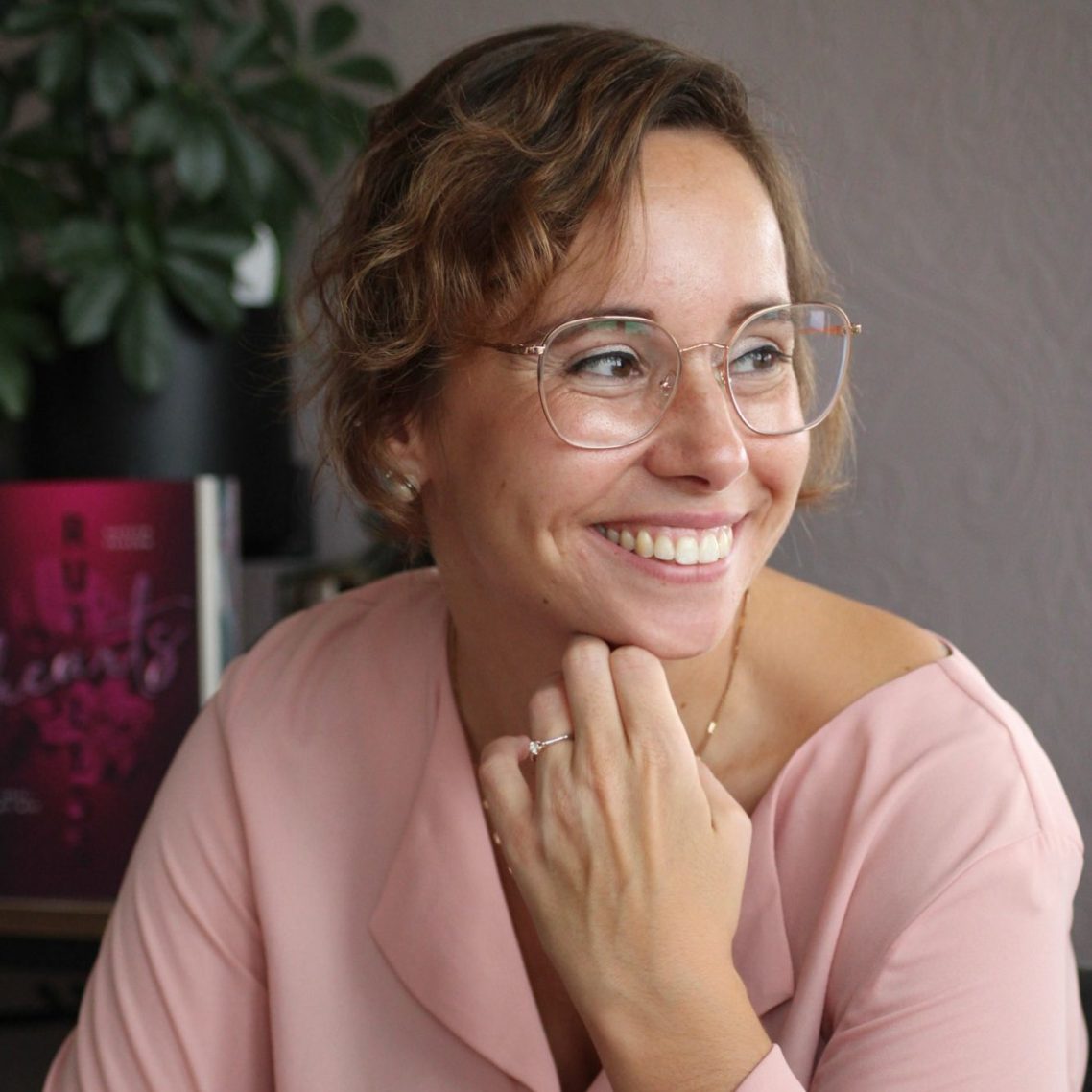 Frau mit lockigem Haar, Brille und rosa Bluse, lächelt freundlich in ein Zimmer.