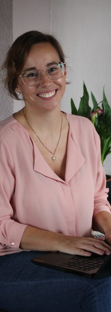 Lächelnde Frau mit Brille, die am Tisch an einem Laptop arbeitet, umgeben von Pflanzen.