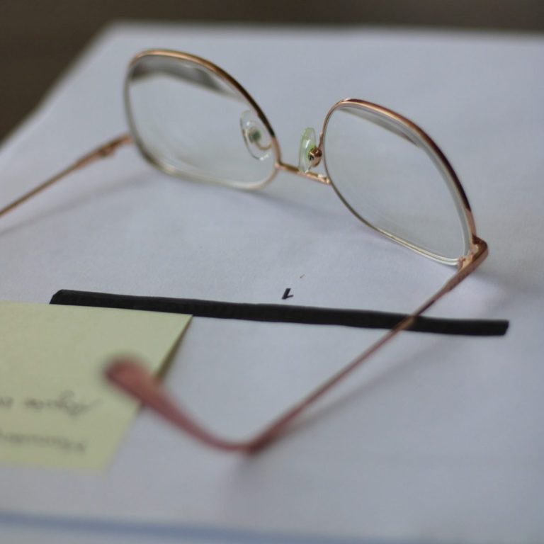 Roséfarbene Brille liegt auf einem Stapel Papier mit einem Post-it-Zettel.
