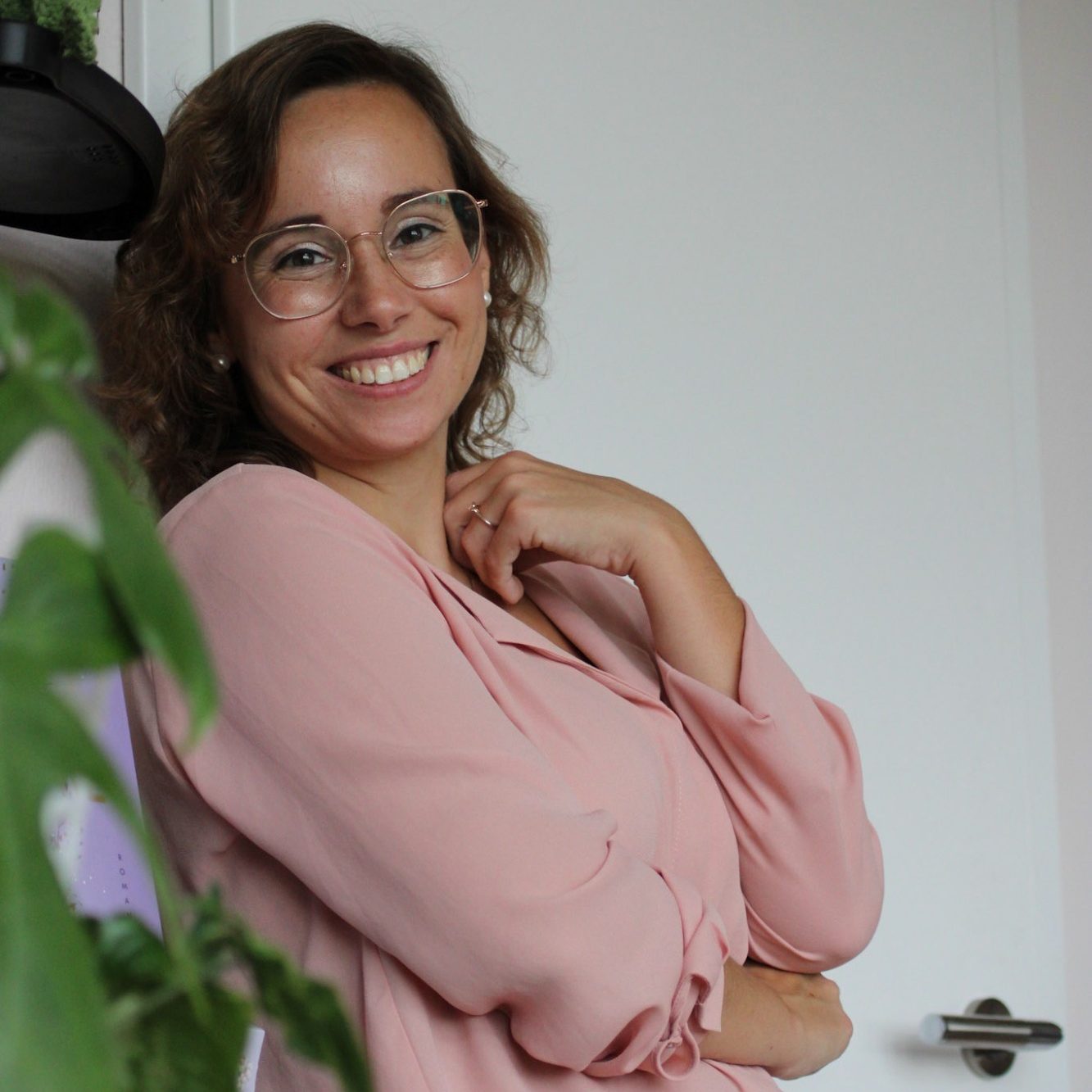 Lächelnde Frau mit Brille und rosa Bluse, steht vor einer Pflanze.