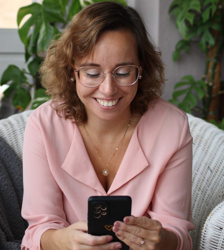 Frau mit Brille in rosa Bluse, lächelnd und auf ein Smartphone schauend.