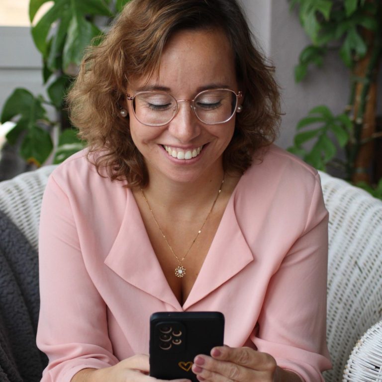 Lächelnde Frau mit Brille, die auf einem Smartphone tippt, umgeben von Pflanzen.